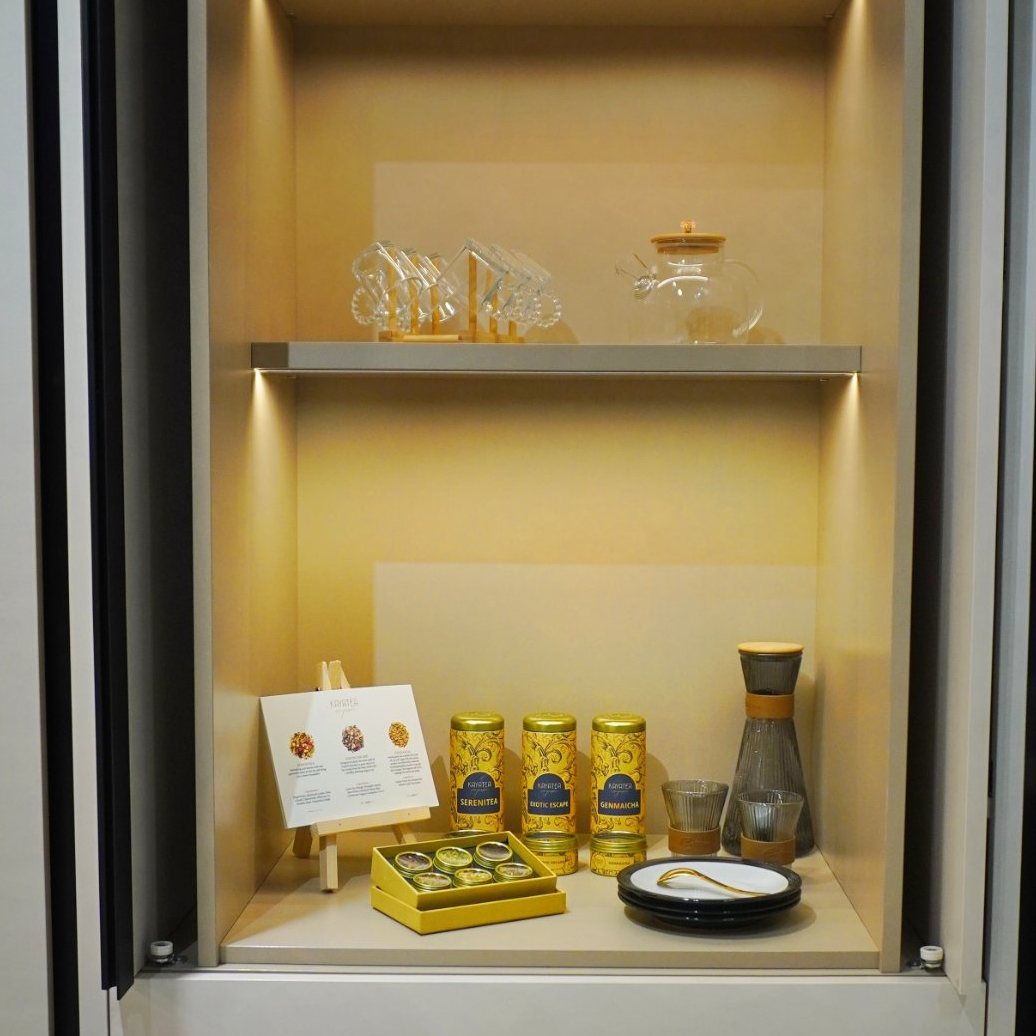 A lit cabinet with Kayatea tins, a Petite Box, glass teaware and a tea card