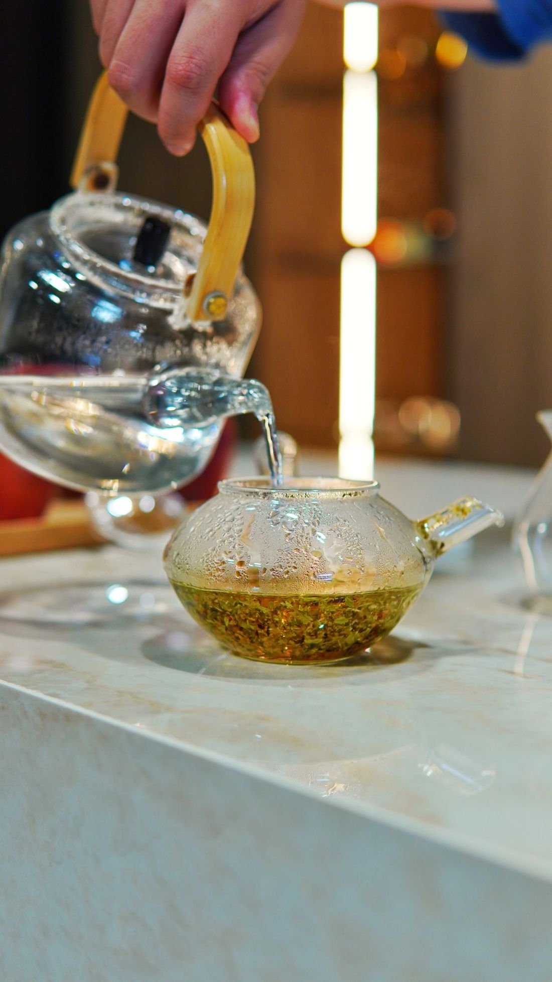 Hot water poured from a glass kettle onto loose leaf tea in a glass teapot