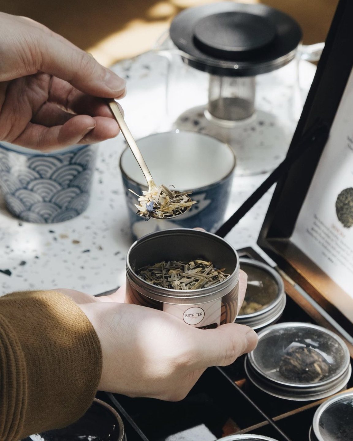 A hand spooning loose leaf tea from a tin, above a wooden box of tea tins and a tasting card