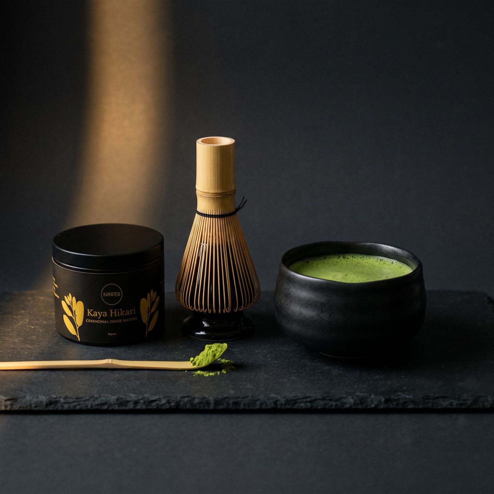 A Kaya Matcha tin, bamboo whisk and a bowl of whisked matcha on dark slate