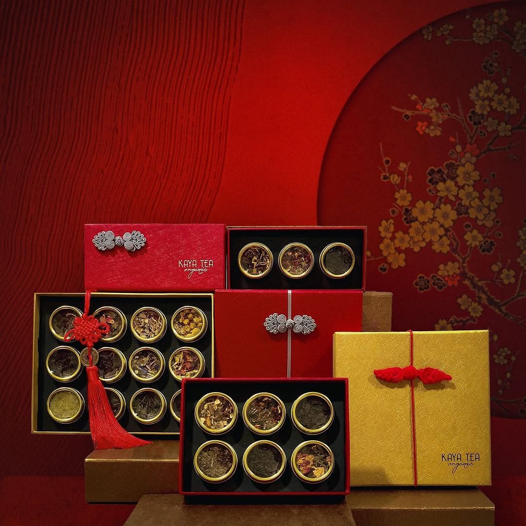 Kayatea Chinese New Year gift boxes in red and gold, with knot closures and a red tassel, against a red plum-blossom backdrop