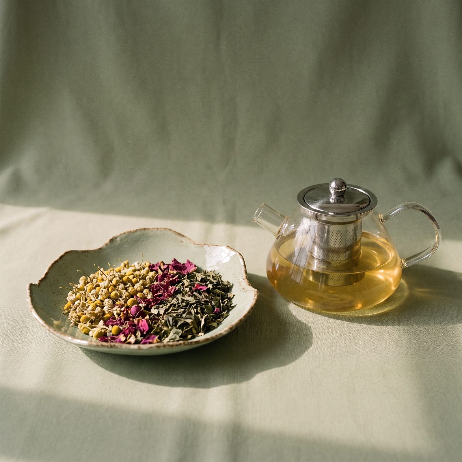 A dish of loose herbal blend beside a glass teapot on sage linen