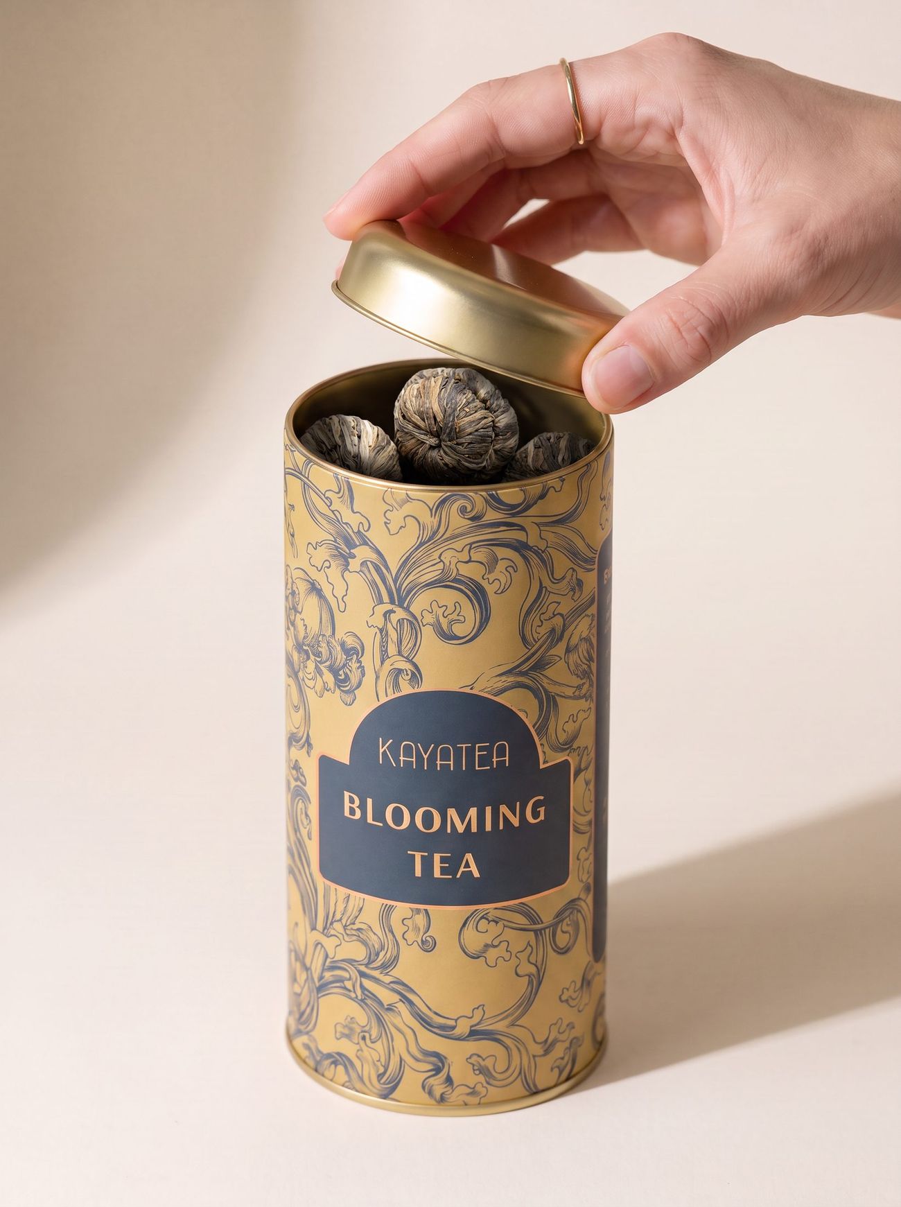 A hand lifting the lid of a Kayatea Blooming Tea tin, the tea bulbs inside